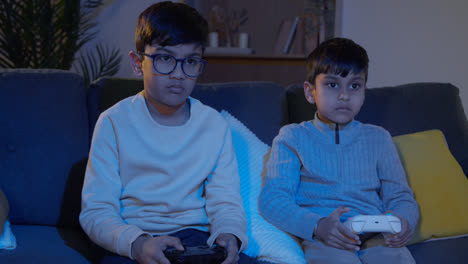 Two-Young-Boys-Sitting-On-Sofa-At-Home-Playing-With-Computer-Games-Console-On-TV-Holding-Controllers-Late-At-Night-2
